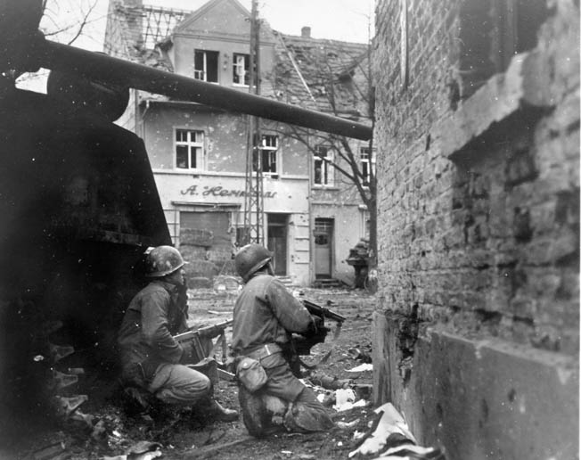 On December 17, 1944, as the German juggernaut rolls forward during the Battle of the Bulge, two infantrymen of the American 83rd Division, both armed with Thompson submachine guns, peer around a corner in the rubble-strewn streets of Gurzenich, Germany.