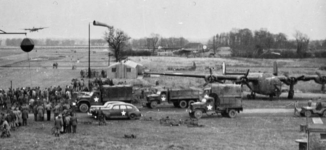 A crowd of onlookers has gathered with its accompanying trucks and staff cars to watch the approach of an overdue Consolidated B-24 Liberator at an airbase in England. The B-24 proved an excellent aircraft in the anti-submarine role and was also one of the primary aircraft deployed during the Allied strategic bombing campaign against Nazi Germany. 