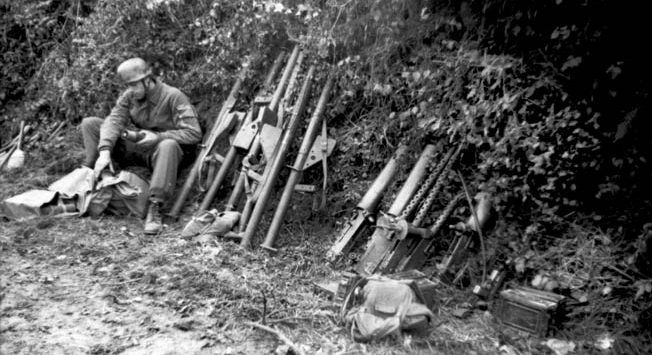 A German paratrooper, sitting in the lee of a hedgerow, prepares ammunition for several shoulder-fired antitank weapons. His arsenal also includes captured American machine guns.
