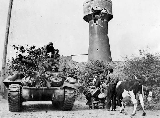 A French family, fleeing the battle area with their cow and meager belongings, watches an American tank destroyer, camouflaged with foliage, rumble by.