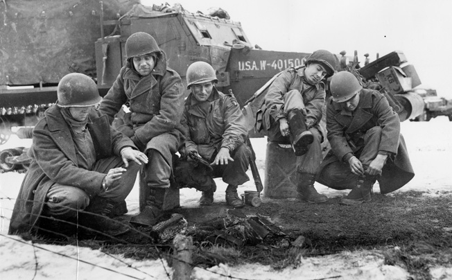 Antiaircraft gun crewmen from the 6th Armored Division and paratroopers from the 101st Airborne warm themselves by a fire while pulling on new overshoes after the relief of Bastogne.