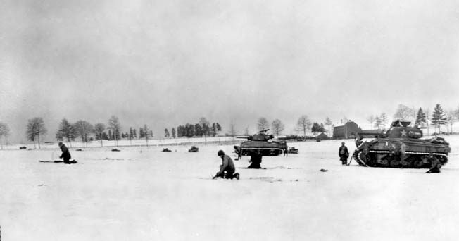 During heavy fighting against German forces in the vicinity of Bastogne, armored infantrymen of the 6th Armored Division advance cautiously across a snow-covered meadow. Sherman tanks are visible in support of the advance.