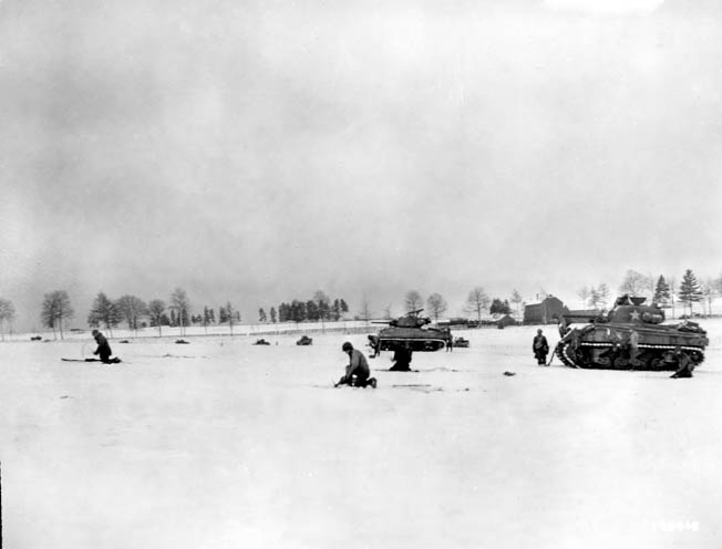 During heavy fighting against German forces in the vicinity of Bastogne, armored infantrymen of the 6th Armored Division advance cautiously across a snow-covered meadow. Sherman tanks are visible in support of the advance.
