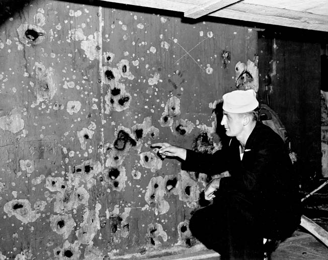 A sailor points to damage sustained by the cruiser USS Boise during the Battle of Cape Esperance. This shredded bulkhead was hit by fragments from a shell that exploded only 30 feet away. This photo was taken during repairs at the Philadelphia Navy Yard.