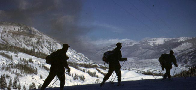 Three 10th Mountain Division soldiers ski above Camp Hale and the Pando Valley. A cloud of dark coal smoke from stoves and passing freight trains darkens the otherwise pristine mountain air.