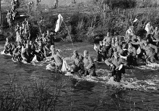 Wading across a small stream and prominently waving white flags, Yugoslav soldiers approach German troops and surrender, preferring captivity to the prospect of combat with an enemy that was already experienced in warfare and confident of victory. During their swift conquest of Yugoslavia, the Germans swept aside most of the organized resistance they encountered.