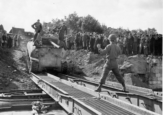 A soldier guides a tank crew onto a Treadway bridge as local villagers look on. Erickson drove his tank across a similar bridge over the Rhine River at night, admitting that if he had seen the bridge in daylight he would have gone AWOL. 