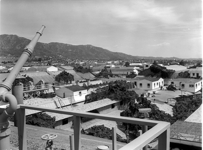 In this view of the Lockheed factory camouflage at Burbank, looking southeasterly toward Glendale, one gets an idea how complex much of the structure really was. On the left in the foreground is a hose nozzle for firefighting. Being made of wood, burlap, and tar, the whole affair was a potential fire hazard.