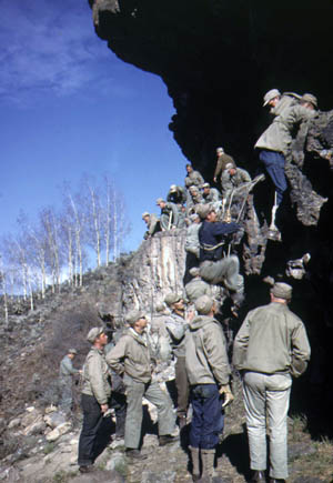 Approximately 20 10th Mountain Division soldiers practice rock climbing at Camp Hale, Colorado. The group is wearing a variety of clothing, including denim jeans and jean jackets.