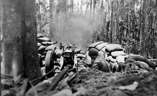 A gunner pulls the lanyard and fires his 75mm pack howitzer through the trees. Smoke from an artillery strike is visible rising from the crest of Hill 260 in the distance. 