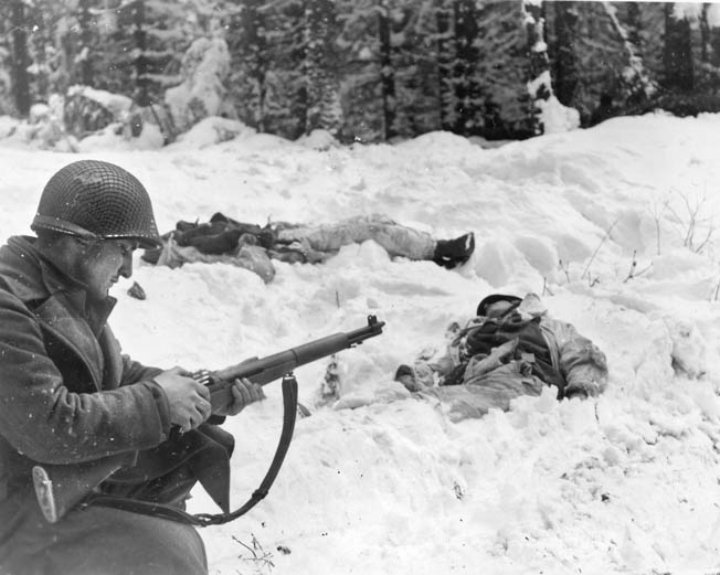 A GI calmly loads his M-1 Garand near two dead Germans whose white camouflage clothing did them no good.