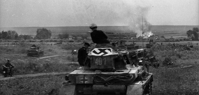 With a Nazi Party flag draped across its turret to show accompanying Luftwaffe planes that these are German troops, columns of tanks roll toward a burning Soviet village. 