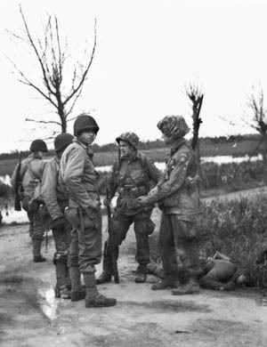 Paratroopers of the 82nd Airborne Division with helmet covers fashioned out of parachute silk chat with 4th Infantry Division soldiers near La Fière while a German soldier lies dead at their feet. Some of the flooded fields are visible in the background.