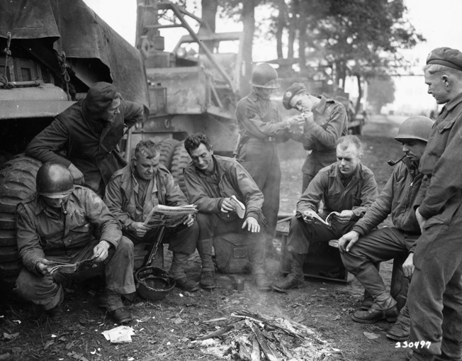 British and American combat engineers swap stories, news, and cigarettes around a small fire as they wait for a call to build a Bailey Bridge near Someren, Holland. The British are with the 15th Scottish Division, while the Americans are with the 7th Armored Division. 