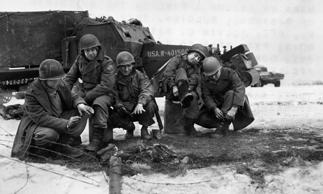 On the outskirts of Bastogne, paratroopers of the 101st Airborne Division and tankers of the 6th Armored Division gather around a fire five days after the surrounded city had been relieved. Some of the men are pulling their newly warmed overshoes over their boots.