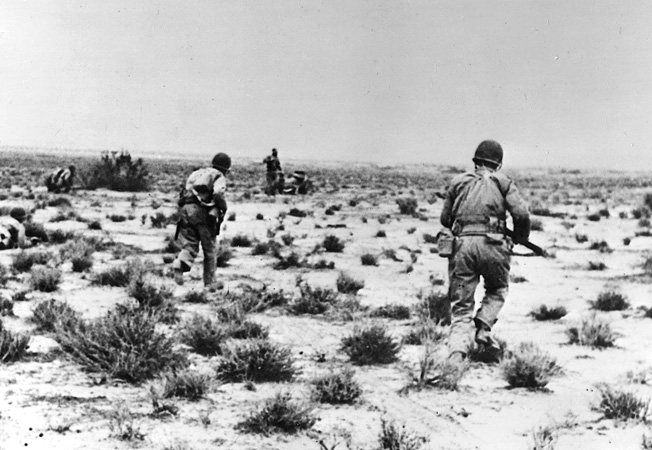 U.S. soldiers, weapons at the ready, advance warily toward German positions across the rock-strewn desert.