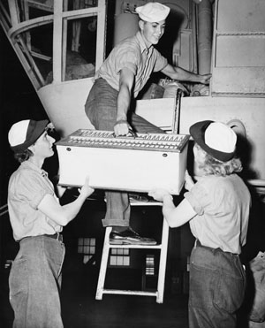 Enclosed in a wood and canvas crate, two birds are taken aloft in a blimp’s gondola. A number of WAVES serving at air stations throughout the U.S. trained pigeons for “air duty.”
