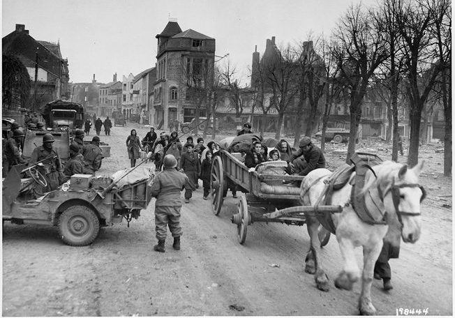 Much of the civilian population of Bastogne left the town with the approach of battle. Here, some of the townspeople, now refugees, seek safety. American troops have halted along the street, where no snow has fallen as of the date of this image.