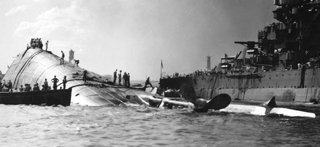 The capsized hull of the battleship USS Oklahoma lies in the shallow water of Pearl Harbor. The Oklahoma was hit by several Japanese torpedoes, causing the ship to capsize and trapping a number of sailors below decks. Feverish efforts to rescue the trapped men were only partially successful.