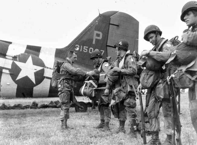 A jumpmaster checks the reserve 'chutes and equipment of paratroopers about to board a C-47. Note that a military censor has obscured the unit patches on the men's left sleeves.