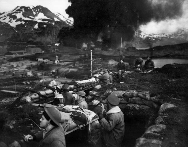 U.S. Marines man their sand-bagged trenches in anticipation of another enemy attack on Dutch Harbor while smoke rises in the background from burning fuel tanks, set afire by a dive bomber. 