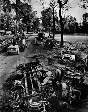 An American half-track towing a trailer speeds past a line of German equipment destroyed by overwhelming Allied air strikes along a road near Ardennes, France. 