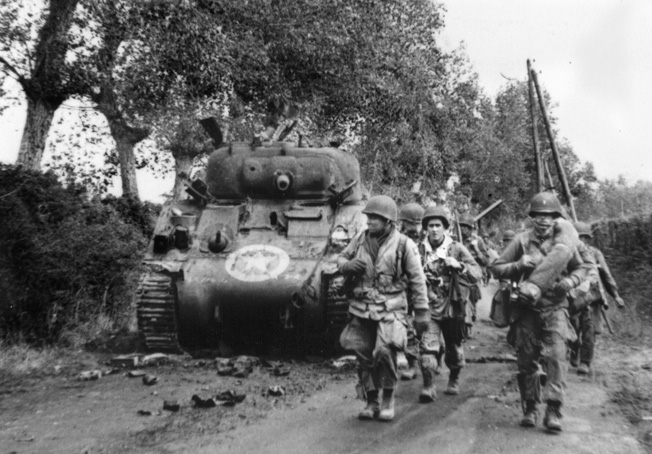 A unit of 82nd Airborne Division troopers advances past a knocked-out M4 Sherman tank along a hedgerow in Normandy.