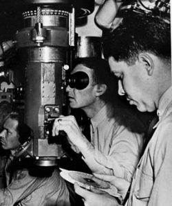A submarine skipper uses the periscope to target an enemy vessel while the executive officer (foreground) uses a manual “Is-Was” slide rule computer to target where the target will be, based on where it was and is. 