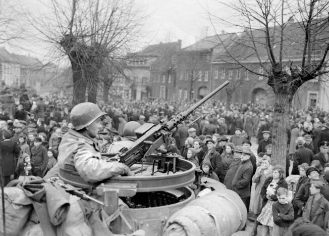An American tanker of the 2nd Armored Division, Ninth Army, sits in the turret of his tank with a .50-caliber machine gun at his disposal. The soldier watches warily as a throng of curious civilians gathers around in the German town of Juchen, captured by American troops in March 1945.