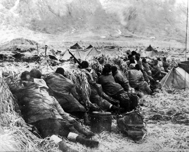 Clad in waterproof clothing to ward off the wet chill of the Aleutians, American troops take cover and scan for enemy snipers located in the mountains above Massacre Bay on Attu Island.