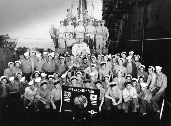 The crew of the USS Archer-Fish poses with the submarine's battle flag, which prominently displays its victories against Japanese naval vessels and merchant shipping. The Archer-Fish was moored in Tokyo Bay alongside the submarine tender USS Proteus when this photograph was taken on September 1, 1945.