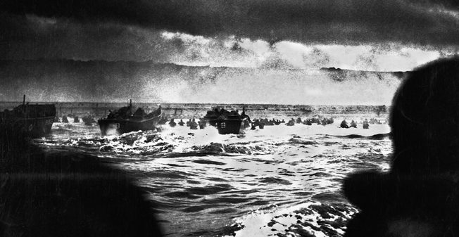 Landing craft churn toward Omaha Beach during the opening minutes of D-Day, June 6, 1944, while some soldiers are seen wading through the chest-high surf.