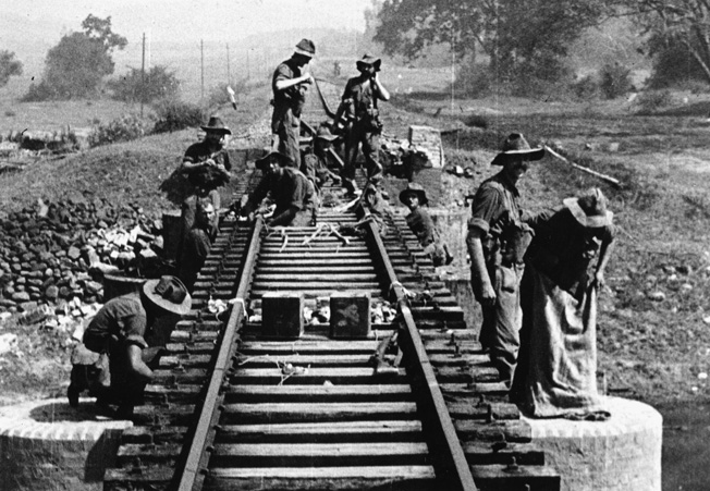 Wearing large-brimmed hats to ward off the tropical sun, engineers of the 77 Brigade wire a section of railroad track with explosives.