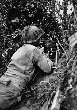 Well hidden by the foliage of a hedgerow in Normandy, an American machine gunner remains on alert during the attack near St. Lô, July 1944. 