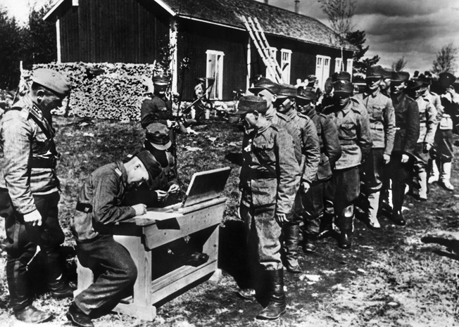 Their nation having succumbed to the Soviet Union during the earlier Winter War, Finnish men line up during the summer of 1941 to join the German Army and continue the fight against the hated communist enemy.