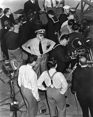 An impatient John Ford watches as a film crew prepares equipment for an upcoming movie scene. Ford pursued an opportunity to assist the American war effort and made the landmark documentary of the Midway fighting in the Pacific.