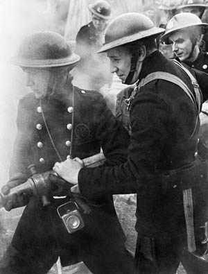 As thick smoke from a raging fire wreaths them, men of the Auxiliary Fire Service attempt to extinguish the blaze after a heavy raid on London during the Blitz.