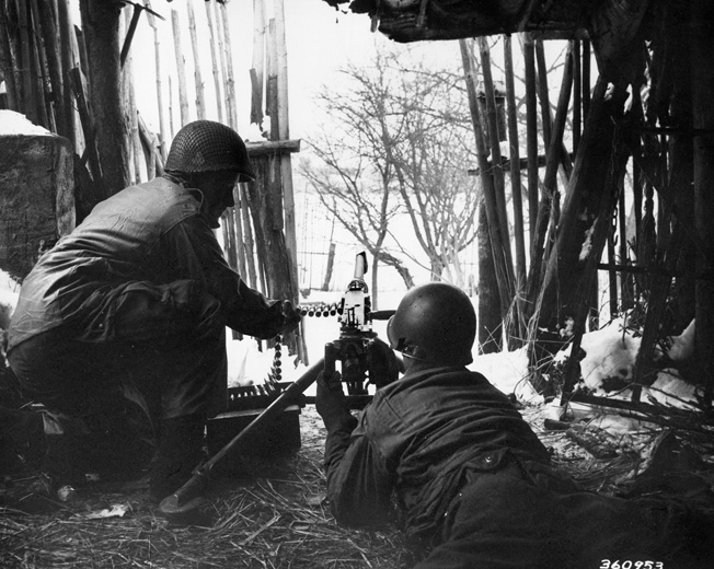 From a position inside a barn near the town of Bastogne, two American soldiers are ready to confront the Germans with their .50-caliber machine gun.