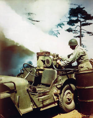 Screening an advancing Army column, an M-2 smoke grenade mounted on a Jeep belches a plume of chemical fog.