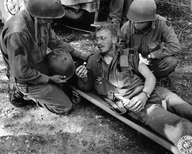 A soldier who has also been wounded in the arm and leg shows a medic the piece of shrapnel that penetrated his M1 helmet but failed to cause a potentially fatal injury.