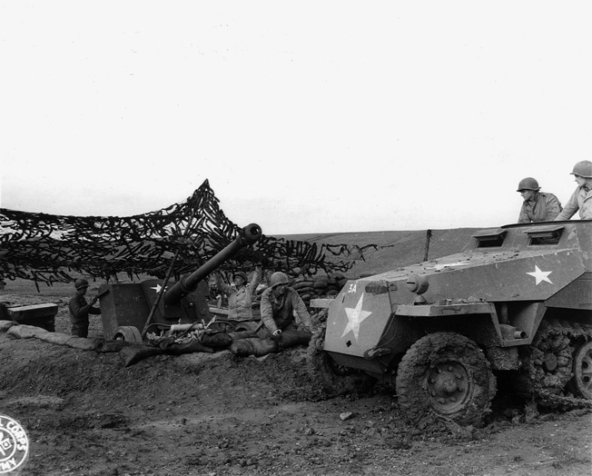 American soldiers prepare a captured German 88mm gun for firing against its former owners while a half-track that originally belonged to the Germans brings ammunition forward.