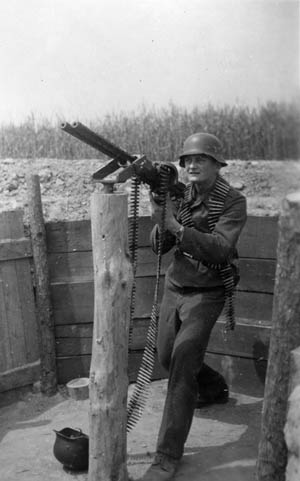 With a belt of ammunition around his neck, Dulias dons a Luftwaffe flak helmet and mans a double-barreled machine gun (MG 81) which has been mounted on a post for gunnery training.