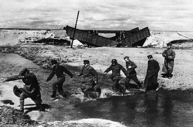 Under the watchful eye of a single Red Army infantryman, a group of German prisoners passes a demolished railway bridge and splashes across a small stream on the Eastern Front.