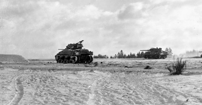 During the Battle of the Bulge in December 1944, an American Sherman tank, accompanied by an M10 tank destroyer, advances steadily across open ground. Such terrain was dangerous, as German antitank teams and hidden guns lurked along the edges of fields and roads.