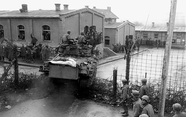 A Sherman tank smashes through the gate of the POW camp at Hammelburg as American and British soldiers wave and celebrate.