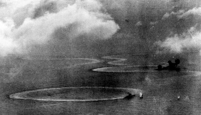 During the Battle of the Philippine Sea, a Japanese heavy cruiser turns in a clockwise circle to evade American dive bombers and torpedo planes. In the distance the plumes of two bomb hits on a Kongo-class battleship are visible, while the stricken vessel narrowly averts a collision with an aircraft carrier also caught in the relentless air attack. 