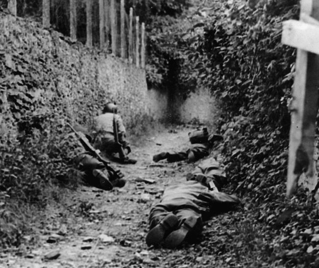 Crouching low against a wall and the eastern mound of a French hedgerow, U.S. soldiers attempt to avoid German artillery fire overhead.