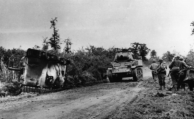 Passing the charred remains of one of its own, a U.S. Stuart light tank makes its way down a dusty road toward St. Lô.