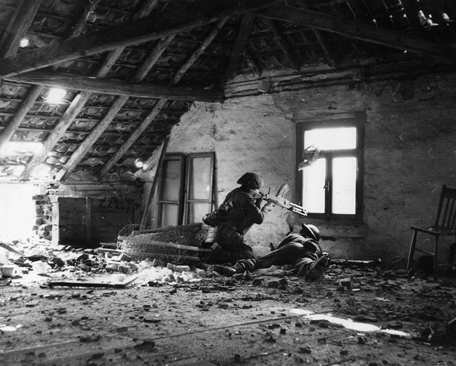 From a window above the streets of a German town, two British soldiers utilizing a Bren gun support comrades below.
