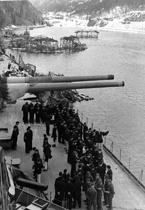 German sailors stand on the deck of the massive battleship Tirpitz moored in a Norwegian fjord sometime between 1942 and 1944. One of the warship’s 15-inch gun turrets is trained abeam, while camouflage floats are seen in the distance. Tirpitz was also well protected by antiaircraft guns on shore.